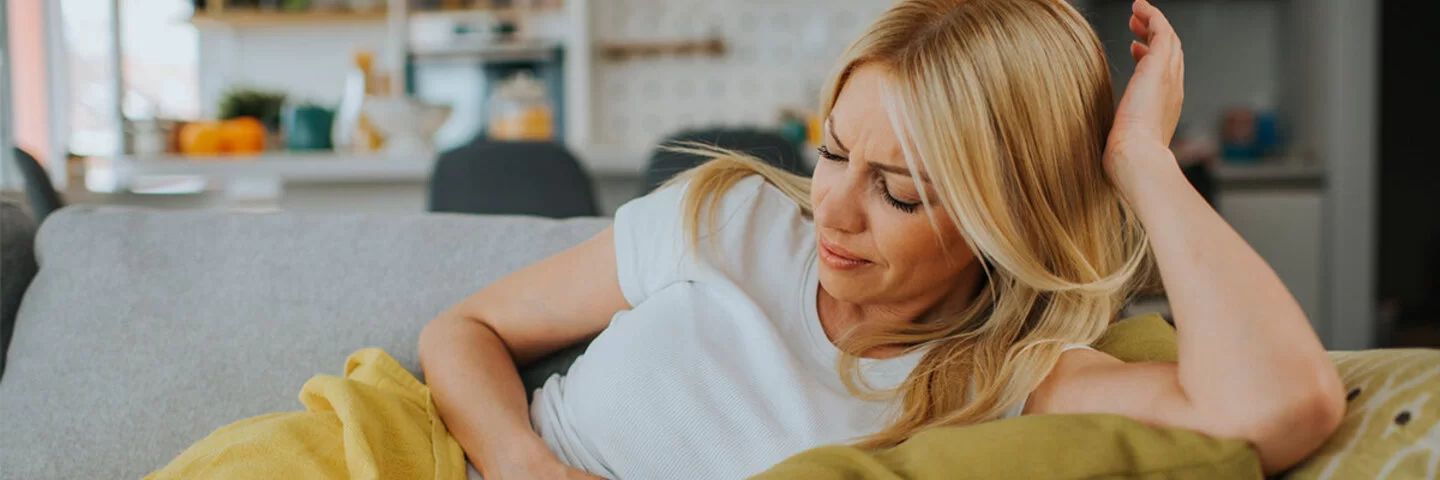 Eine Frau liegt mit schmerzverzerrtem Gesichtsausdruck auf dem Sofa und hält sich den Bauch.