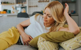 Eine Frau liegt mit schmerzverzerrtem Gesichtsausdruck auf dem Sofa und hält sich den Bauch.