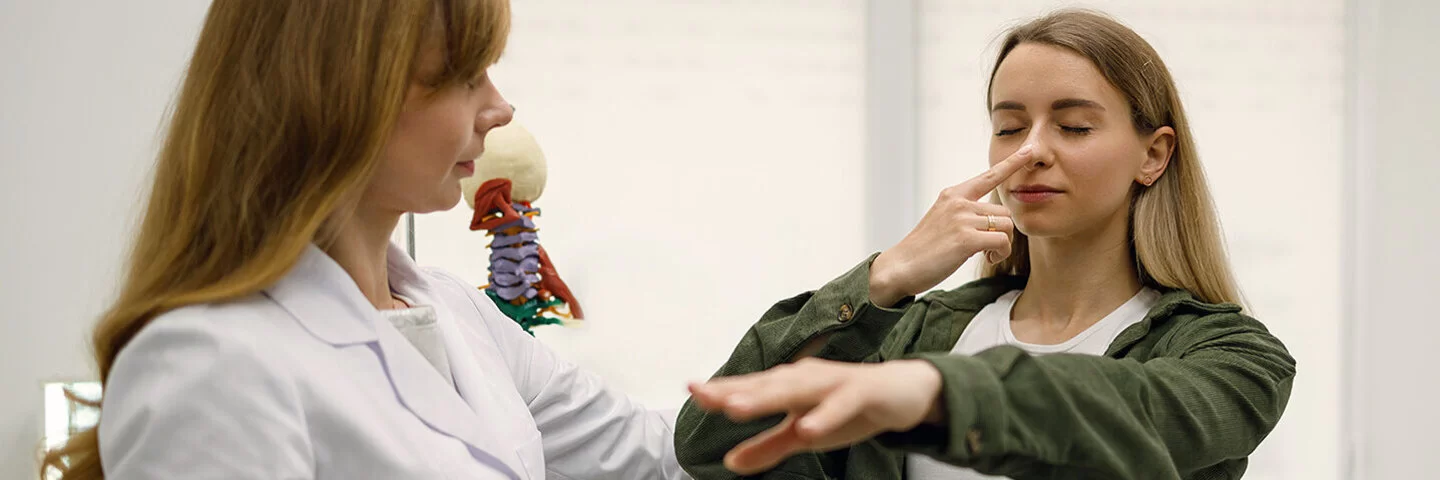 Eine junge Frau steht neben einer Neurologin. Sie hat die Augen geschlossen und hält ihren linken Arm ausgestreckt, während sie mit ihrem rechten Zeigefinger ihre Nasenspitze berührt. Die Szene deutet auf einen neurologischen Test hin.