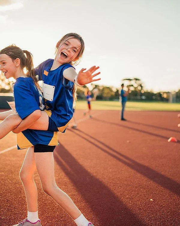 Ein Mädchen trägt ein anderes Mädchen, das lachend in die Kamera winkt, auf ihrem Rücken. Beide tragen blaue Trikots. Im Hintergrund ist ein großer roter Sportplatz bei Sonnenuntergang mit anderen Kindern zu sehen.