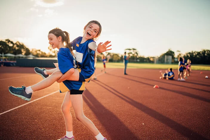 Ein Mädchen trägt ein anderes Mädchen, das lachend in die Kamera winkt, auf ihrem Rücken. Beide tragen blaue Trikots. Im Hintergrund ist ein großer roter Sportplatz bei Sonnenuntergang mit anderen Kindern zu sehen.