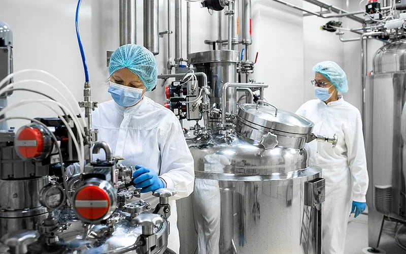 In einem Labor stehen zwei Mitarbeiterinnen in Schutzkleidung und mit OP-Masken an Apparaturen mit Kesseln, Ventilen und Schläuchen.