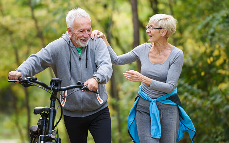 Eine Frau und ein Mann, der ein Fahrrad schiebt, gehen im Wald spazieren und unterhalten sich.