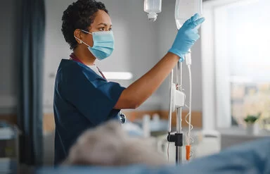Eine Krankenschwester überprüft konzentriert den Tropf eines Patienten. Sie trägt einen Mundschutz und Schutzkleidung, während sie ihre klinische Arbeit verrichtet.