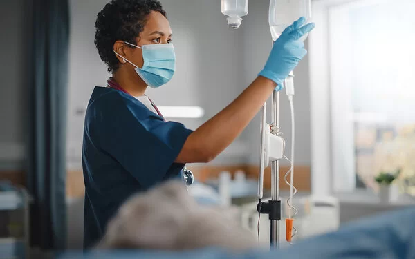 Eine Krankenschwester überprüft konzentriert den Tropf eines Patienten. Sie trägt einen Mundschutz und Schutzkleidung, während sie ihre klinische Arbeit verrichtet.