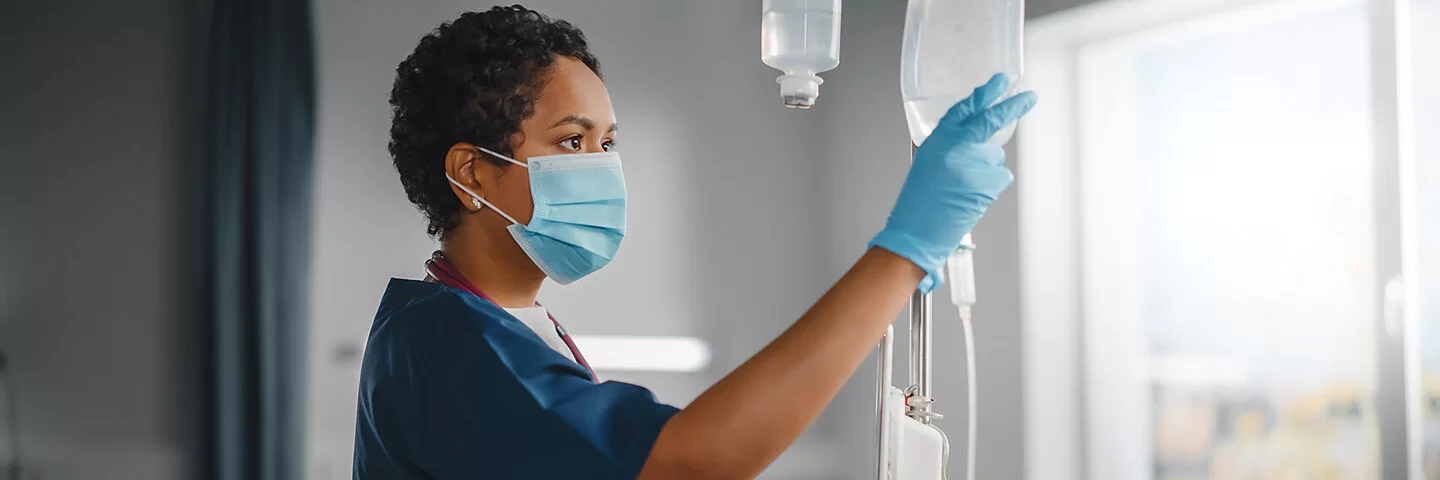 Eine Krankenschwester überprüft konzentriert den Tropf eines Patienten. Sie trägt einen Mundschutz und Schutzkleidung, während sie ihre klinische Arbeit verrichtet.