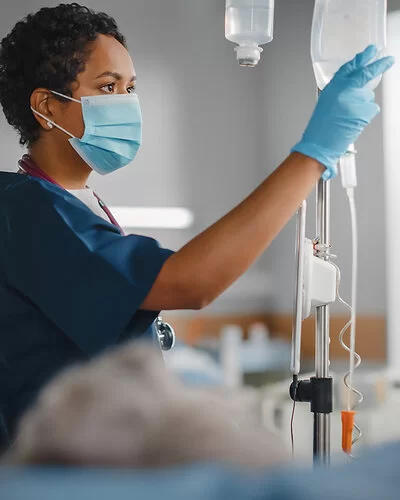 Eine Krankenschwester überprüft konzentriert den Tropf eines Patienten. Sie trägt einen Mundschutz und Schutzkleidung, während sie ihre klinische Arbeit verrichtet.