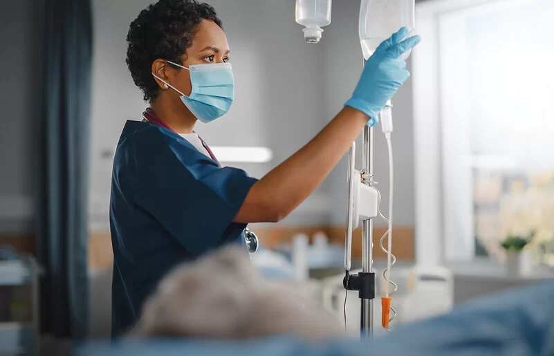 Eine Krankenschwester überprüft konzentriert den Tropf eines Patienten. Sie trägt einen Mundschutz und Schutzkleidung, während sie ihre klinische Arbeit verrichtet.