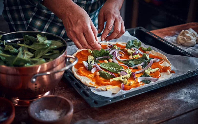 Eine Person belegt eine Pizza als gesundes Fast Food mit reichlich Gemüse.