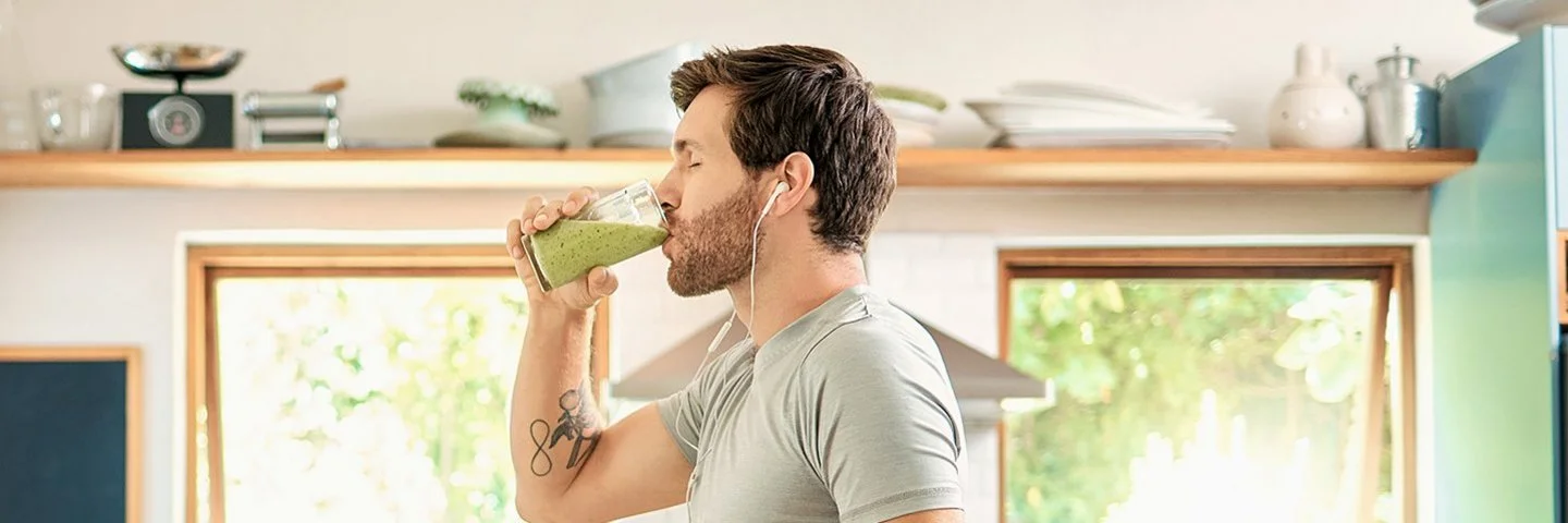 Ein sportlicher junger Mann mit Kopfhörern in den Ohren genießt mit geschlossenen Augen in seiner Küche ein Glas gesunden grünen Detox-Smoothie.