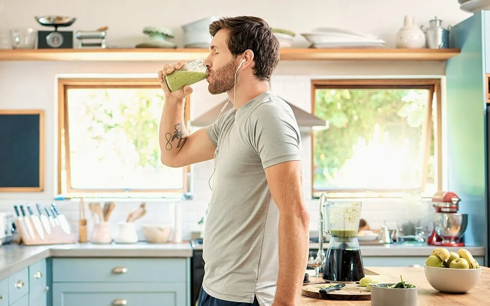 Ein sportlicher junger Mann mit Kopfhörern in den Ohren genießt mit geschlossenen Augen in seiner Küche ein Glas gesunden grünen Detox-Smoothie.