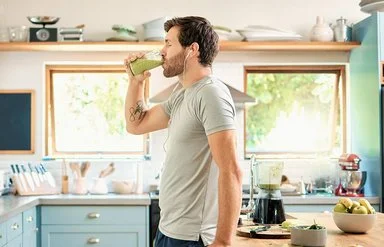 Ein sportlicher junger Mann mit Kopfhörern in den Ohren genießt mit geschlossenen Augen in seiner Küche ein Glas gesunden grünen Detox-Smoothie.