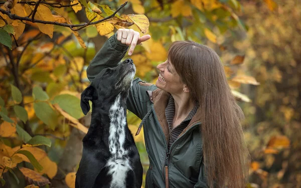 Melanie von Zelewski schaut lachend ihren großen, schwarzen Hund an, der neben ihr im herbstlichen Wald sitzt.