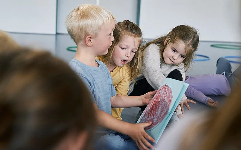 Nahaufnahme von drei Kindern, die auf einem blauen Hallenboden sitzen. Ein blonder Junge in einem hellblauen T-Shirt hält ein blaues Blatt Papier mit einem aufgemalten roten Kreis in den Händen. Die beiden Mädchen neben ihm schauen neugierig auf das Papier.