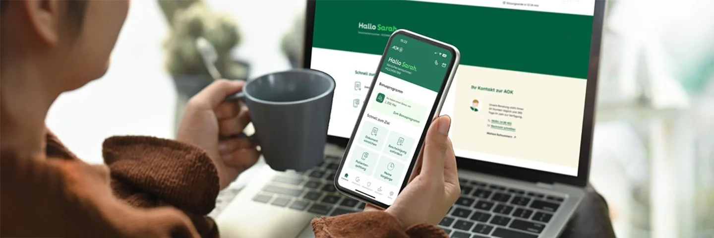 Eine Frau sitzt vor ihrem Laptop und hält in der einen Hand eine Kaffeetasse und in der anderen ein Handy. Auf beiden Bildschirmen ist die App Meine AOK zu sehen.