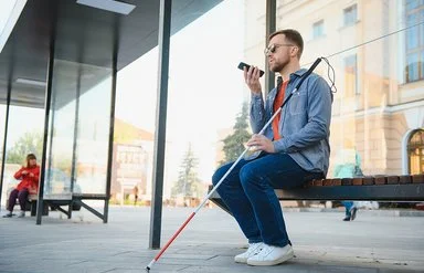 Ein junger Mann mit dunkler Sonnenbrille sitzt auf einer Bank. In der linken Hand hält er einen Blindenstock, in der rechten ein Smartphone.