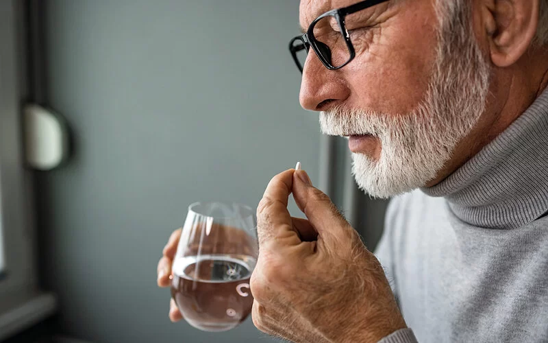 Ein alter Mann nimmt eine Tablette mit einem Schluck Wasser aus einem Glas ein.