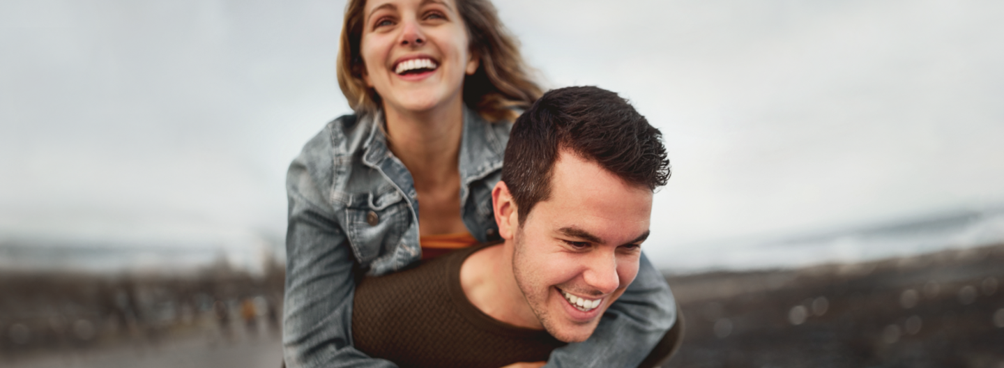 Ein junger Mann trägt eine Frau auf seinem Rücken. Beide lachen.