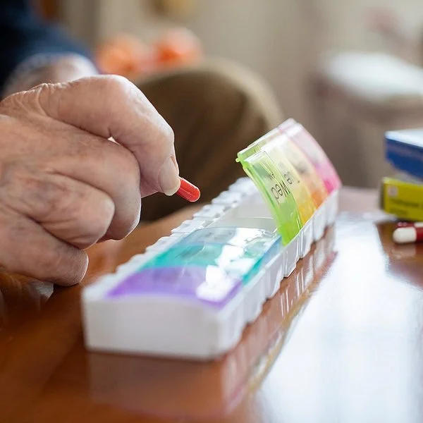 Auf einem Tisch liegen mehrere Tablettenschachteln, Pillengläser und eine Tablettenbox. Die Fächer für die einzelnen Tage haben eigene Deckel, von denen vier geöffnet sind. Die Hand eines älteren Menschen befüllt die Box mit einer Kapsel.