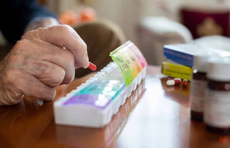 Auf einem Tisch liegen mehrere Tablettenschachteln, Pillengläser und eine Tablettenbox. Die Fächer für die einzelnen Tage haben eigene Deckel, von denen vier geöffnet sind. Die Hand eines älteren Menschen befüllt die Box mit einer Kapsel.