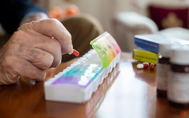 Auf einem Tisch liegen mehrere Tablettenschachteln, Pillengläser und eine Tablettenbox. Die Fächer für die einzelnen Tage haben eigene Deckel, von denen vier geöffnet sind. Die Hand eines älteren Menschen befüllt die Box mit einer Kapsel.