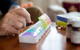 Auf einem Tisch liegen mehrere Tablettenschachteln, Pillengläser und eine Tablettenbox. Die Fächer für die einzelnen Tage haben eigene Deckel, von denen vier geöffnet sind. Die Hand eines älteren Menschen befüllt die Box mit einer Kapsel.