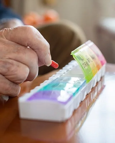Auf einem Tisch liegen mehrere Tablettenschachteln, Pillengläser und eine Tablettenbox. Die Fächer für die einzelnen Tage haben eigene Deckel, von denen vier geöffnet sind. Die Hand eines älteren Menschen befüllt die Box mit einer Kapsel.