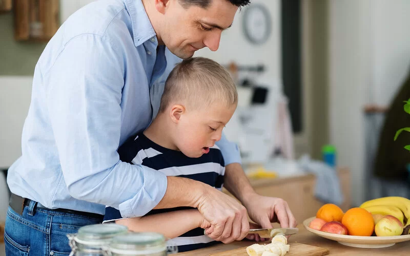 Ein Mann steht hinter einem Jungen mit Trisomie 21 und hilft ihm beim Schneiden einer Banane, indem er die Hände des Jungen führt.