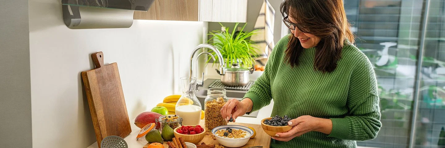 Frau in grünem Pullover bereitet in einer modernen Küche eine Schüssel Müsli mit Heidelbeeren zu. Auf der Arbeitsfläche stehen Milch, Obst, Beeren, Nüsse und Zimtstangen.