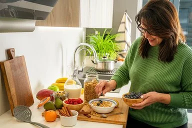 Frau in grünem Pullover bereitet in einer modernen Küche eine Schüssel Müsli mit Heidelbeeren zu. Auf der Arbeitsfläche stehen Milch, Obst, Beeren, Nüsse und Zimtstangen.