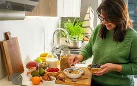 Frau in grünem Pullover bereitet in einer modernen Küche eine Schüssel Müsli mit Heidelbeeren zu. Auf der Arbeitsfläche stehen Milch, Obst, Beeren, Nüsse und Zimtstangen.