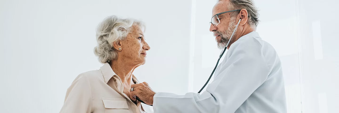 Ein älterer Arzt hört bei einer Seniorin, die auf einer Untersuchungsliege sitzt, das Herz ab.