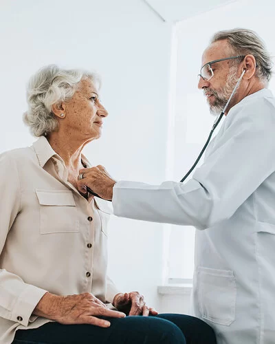 Ein älterer Arzt hört bei einer Seniorin, die auf einer Untersuchungsliege sitzt, das Herz ab.