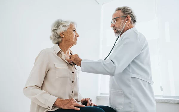 Ein älterer Arzt hört bei einer Seniorin, die auf einer Untersuchungsliege sitzt, das Herz ab.