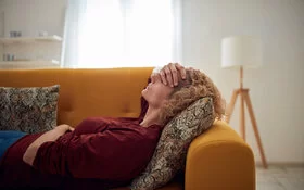 Frau liegt auf einer Couch mit ihrer Hand auf der Stirn, mit einem Arm hält sie sich ihren Bauch.