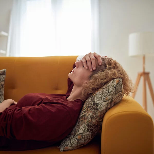 Frau liegt auf einer Couch mit ihrer Hand auf der Stirn, mit einem Arm hält sie sich ihren Bauch.