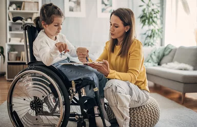 Ein Mädchen sitzt im Rollstuhl. Die Mutter sitzt auf einem Sitzkissen vor der Tochter und hilft dem Mädchen, einen Stift über eine Fläche zu führen.