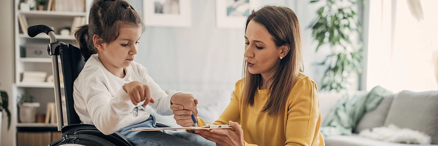 Ein Mädchen sitzt im Rollstuhl. Die Mutter sitzt auf einem Sitzkissen vor der Tochter und hilft dem Mädchen, einen Stift über eine Fläche zu führen.