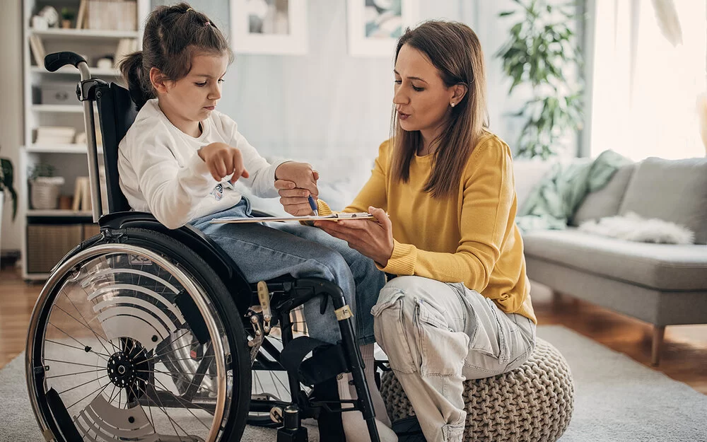 Ein Mädchen sitzt im Rollstuhl. Die Mutter sitzt auf einem Sitzkissen vor der Tochter und hilft dem Mädchen, einen Stift über eine Fläche zu führen.