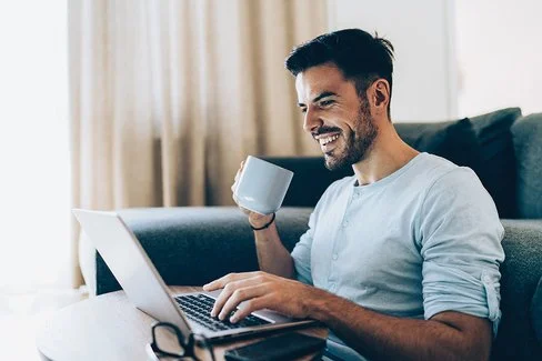 Ein Mann sitzt auf einem Sofa mit einem Laptop und einer Tasse Kaffee in der Hand.