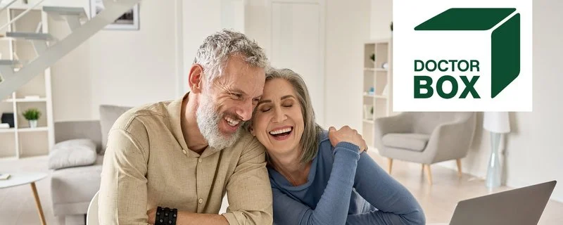 Glückliches, reifes Ehepaar im besten Alter, das lachend und eng verbunden am Tisch zu Hause mit einem Laptop sitzt.