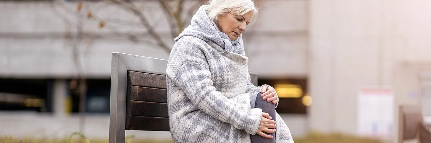Eine Frau im mittleren Alter sitzt auf einer Bank in einer Grünanlage. Sie trägt einen dicken Wintermantel und reibt sich das Knie.