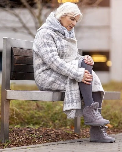 Eine Frau im mittleren Alter sitzt auf einer Bank in einer Grünanlage. Sie trägt einen dicken Wintermantel und reibt sich das Knie.