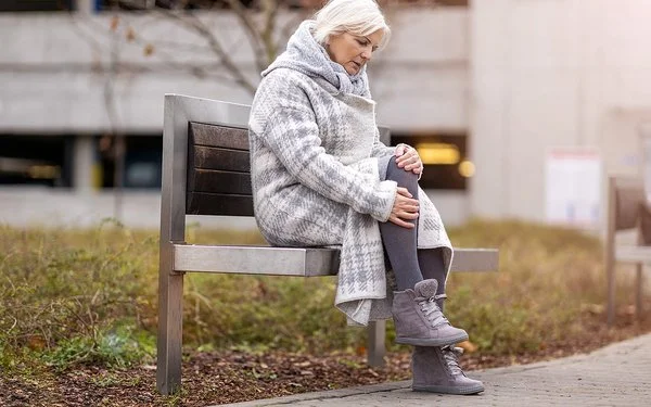 Eine Frau im mittleren Alter sitzt auf einer Bank in einer Grünanlage. Sie trägt einen dicken Wintermantel und reibt sich das Knie.