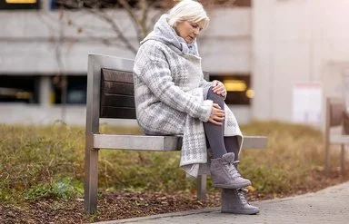 Eine Frau im mittleren Alter sitzt auf einer Bank in einer Grünanlage. Sie trägt einen dicken Wintermantel und reibt sich das Knie.