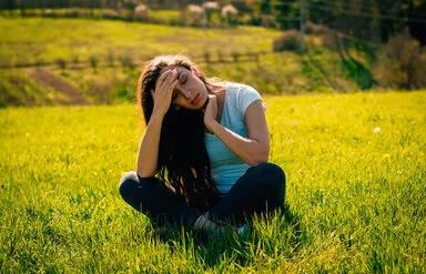 Eine junge braunhaarige Frau sitzt im Schneidersitz auf einem Feld. Sie hält sich mit schmerzvollem Blick die Hand an die Stirn.