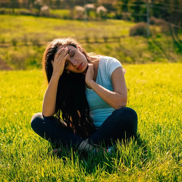Eine junge braunhaarige Frau sitzt im Schneidersitz auf einem Feld. Sie hält sich mit schmerzvollem Blick die Hand an die Stirn.