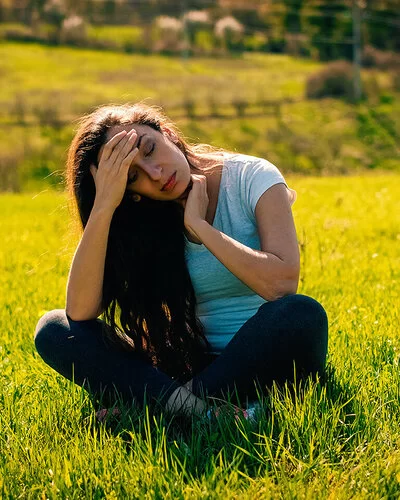 Eine junge braunhaarige Frau sitzt im Schneidersitz auf einem Feld. Sie hält sich mit schmerzvollem Blick die Hand an die Stirn.