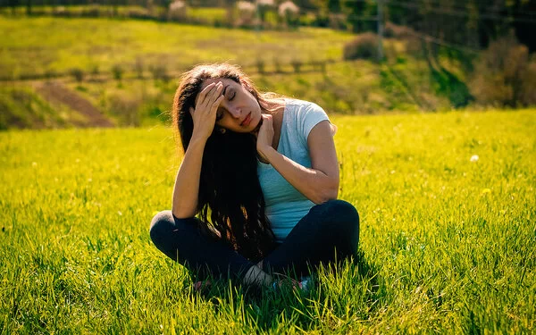 Eine junge braunhaarige Frau sitzt im Schneidersitz auf einem Feld. Sie hält sich mit schmerzvollem Blick die Hand an die Stirn.