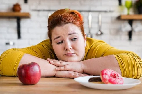 Eine etwas fülligere Frau sitzt mit verschränkten Armen an einem Tisch. Die Frau kann sich nicht zwischen einem Apfel oder einem Donut entscheiden, die vor ihr stehen.